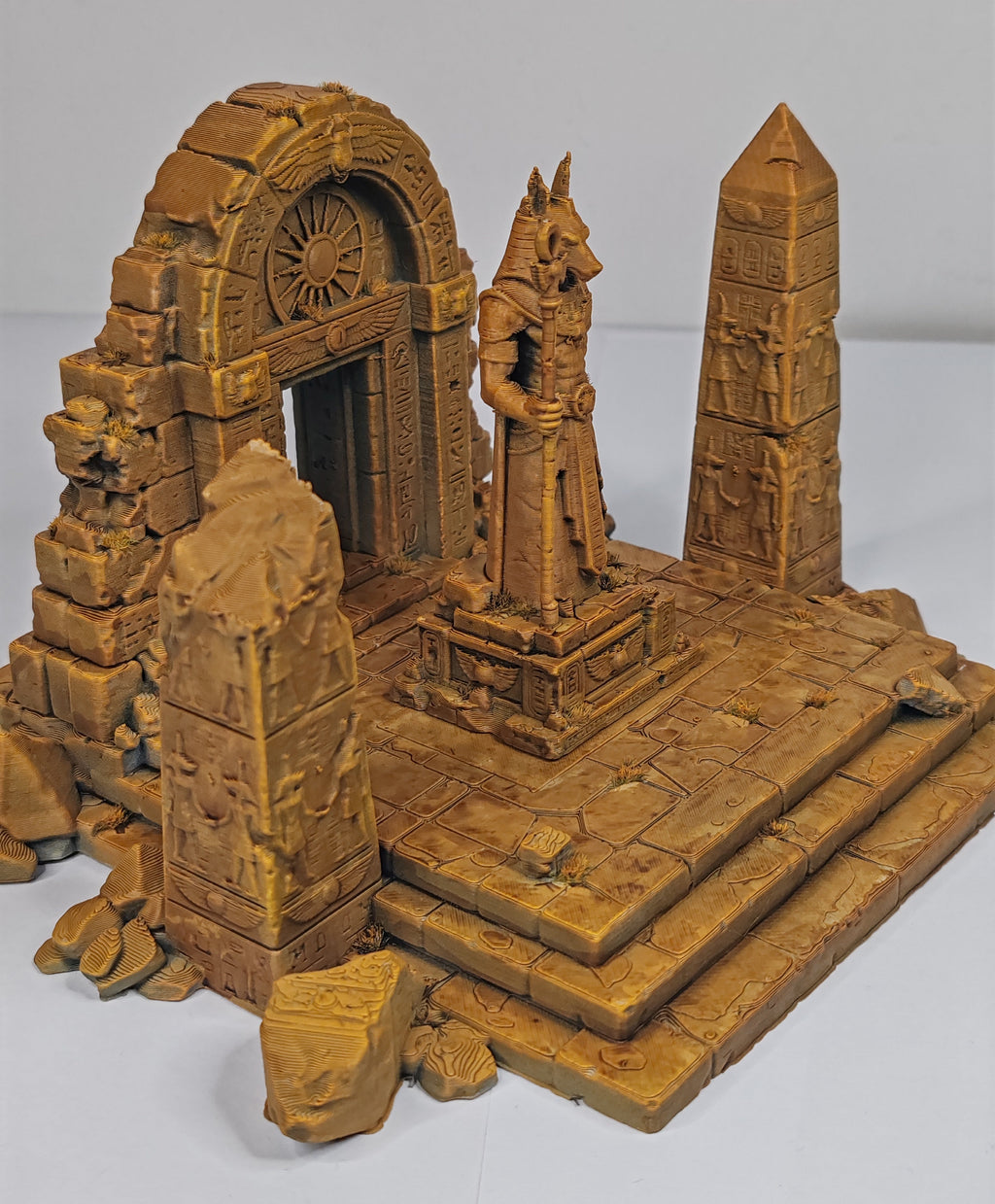 A miniature fantasy ruins model featuring a stone archway with a carved doorway, a central statue on a pedestal, a tall obelisk, and tiered steps, shown on a plain light background.