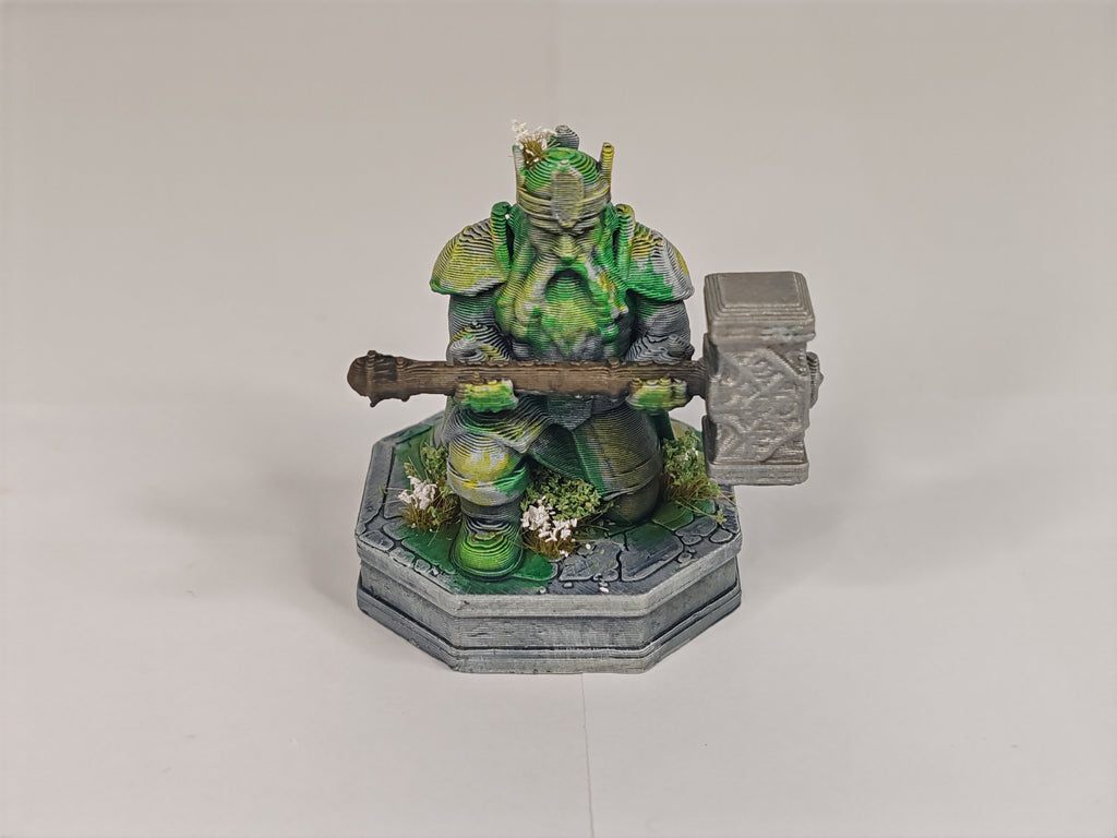A painted fantasy miniature figurine of a green-skinned warrior kneeling and holding a large gray mace, mounted on a gray stone-like base with small white flower details, photographed against a plain light gray studio background.