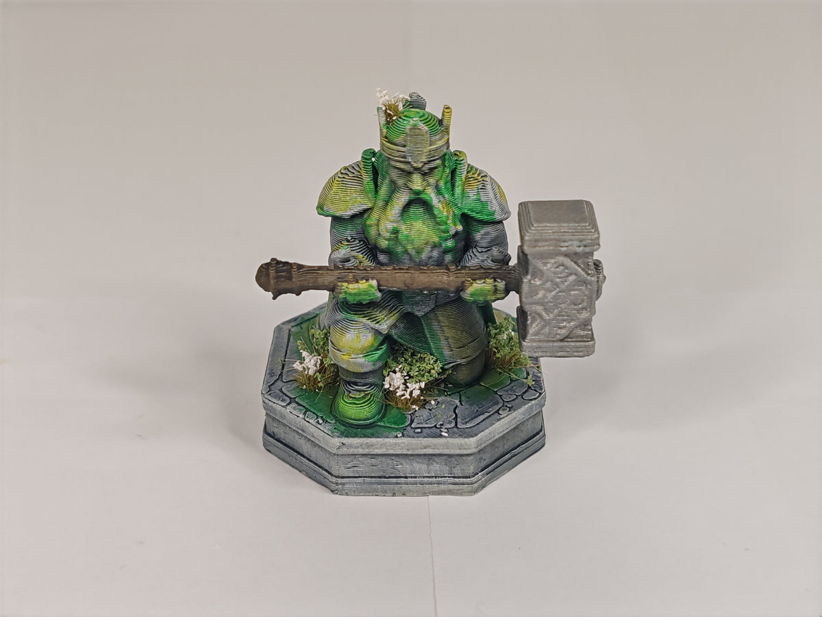 A painted fantasy miniature figurine of a green-skinned warrior kneeling and holding a large gray mace, mounted on a gray stone-like base with small white flower details, photographed against a plain light gray studio background.