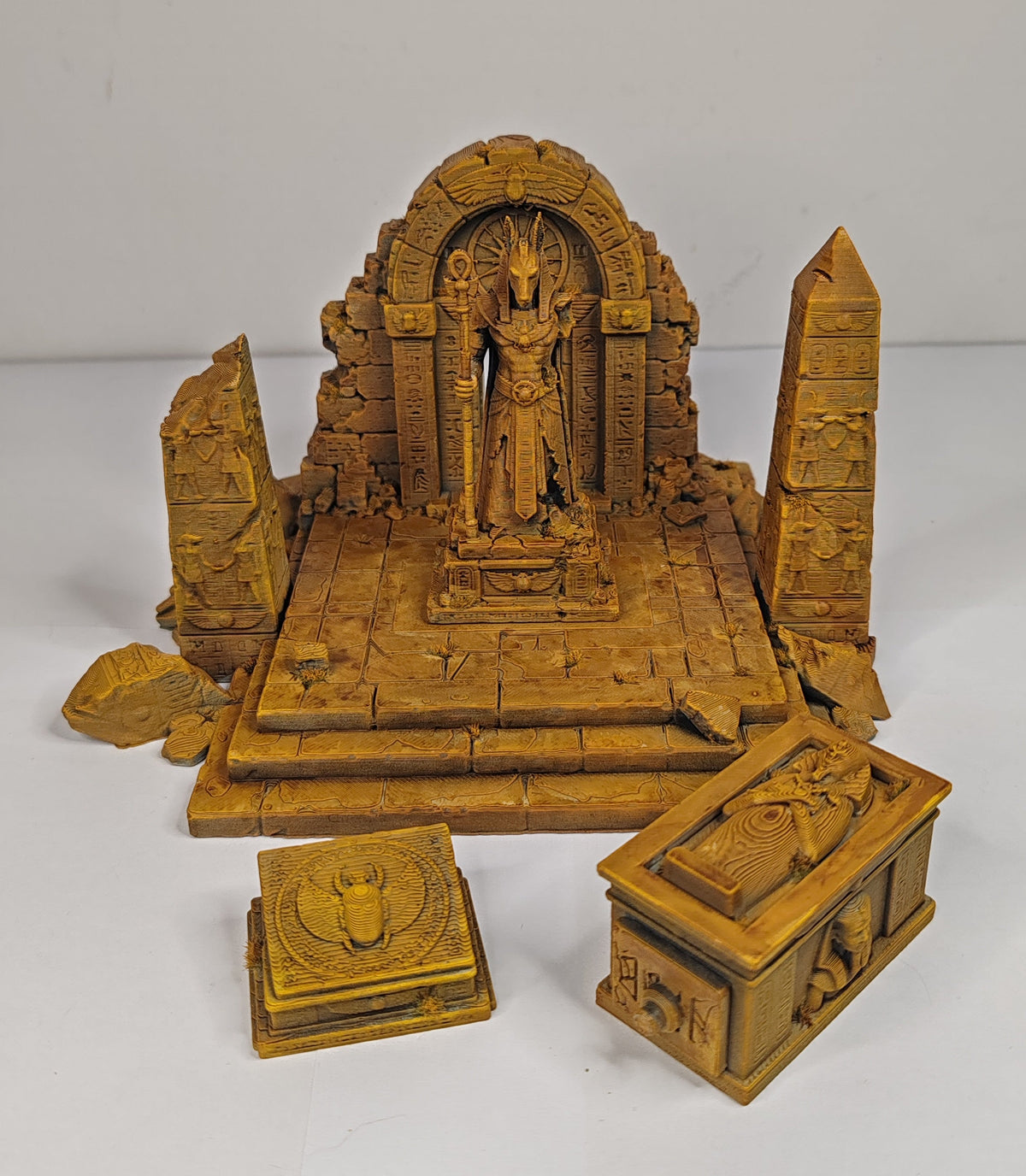 A carved wooden miniature temple display featuring a central deity statue under an arched backdrop, flanked by two obelisks, with two small decorative boxes in front, all in a warm golden-brown finish on a white studio background.
