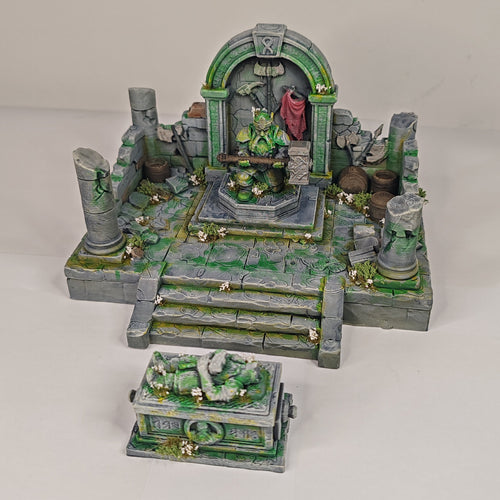 A miniature fantasy ruins set with a stone archway, steps, pillars, and a small chest or crate in front, photographed on a plain light background.