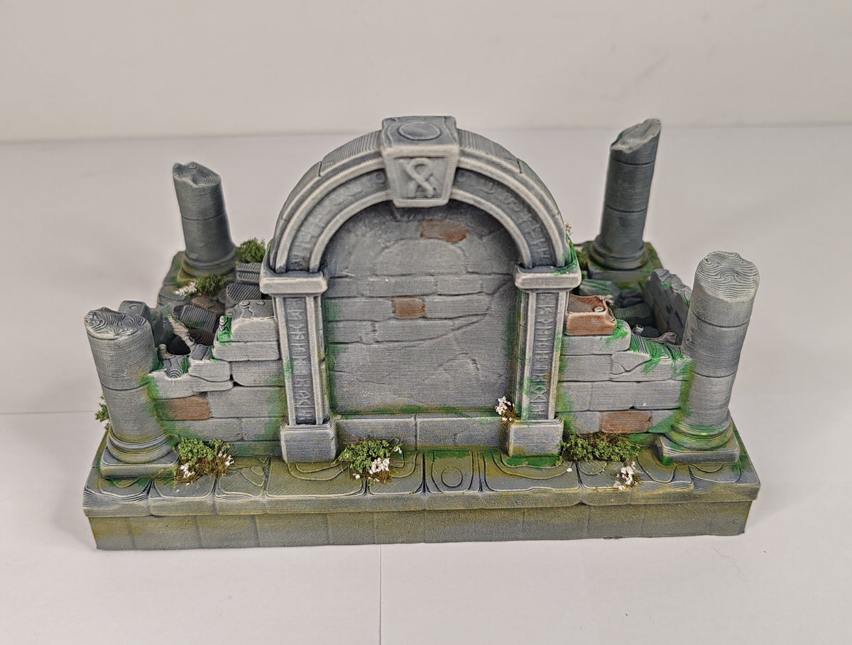 A miniature gray stone ruin tabletop terrain piece with an arched doorway, broken columns, and green moss details, photographed on a plain white background.
