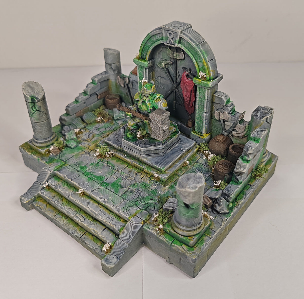 A painted miniature tabletop terrain piece depicting a ruined stone structure with green moss-like detailing, gray pillars, stairs, and a central archway, photographed on a plain light background.