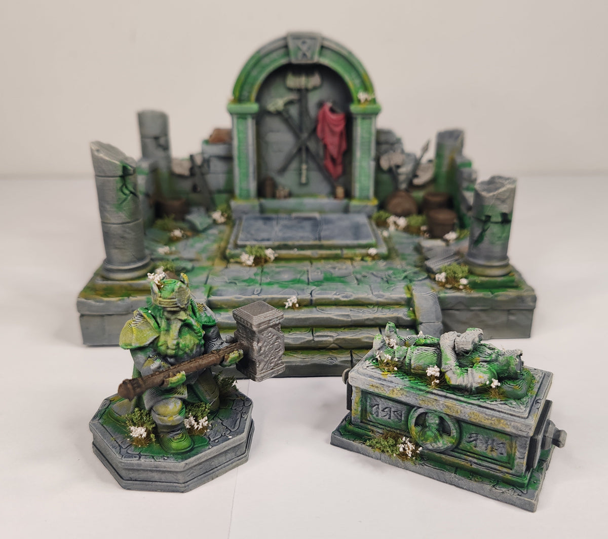 A painted tabletop miniature terrain set featuring a green stone tomb chamber with a red draped banner, a green-armored warrior holding a mace on a base, and a matching green stone chest or coffin piece in front, photographed on a plain white studio background.