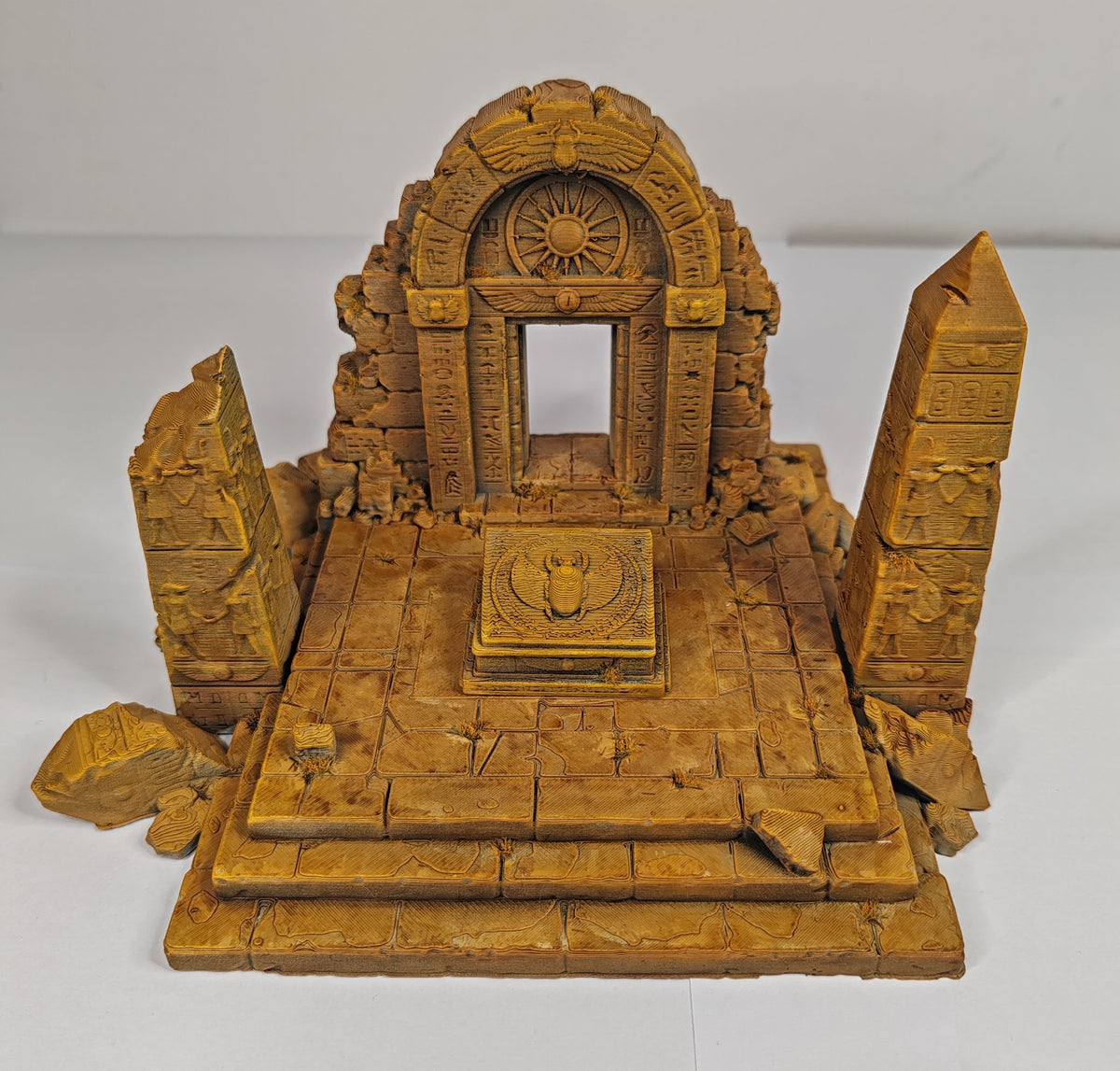 A miniature golden-brown fantasy ruins model, featuring an arched doorway with a sun emblem, a central pedestal, and two tall broken obelisks on a tiled platform, set against a plain white background.