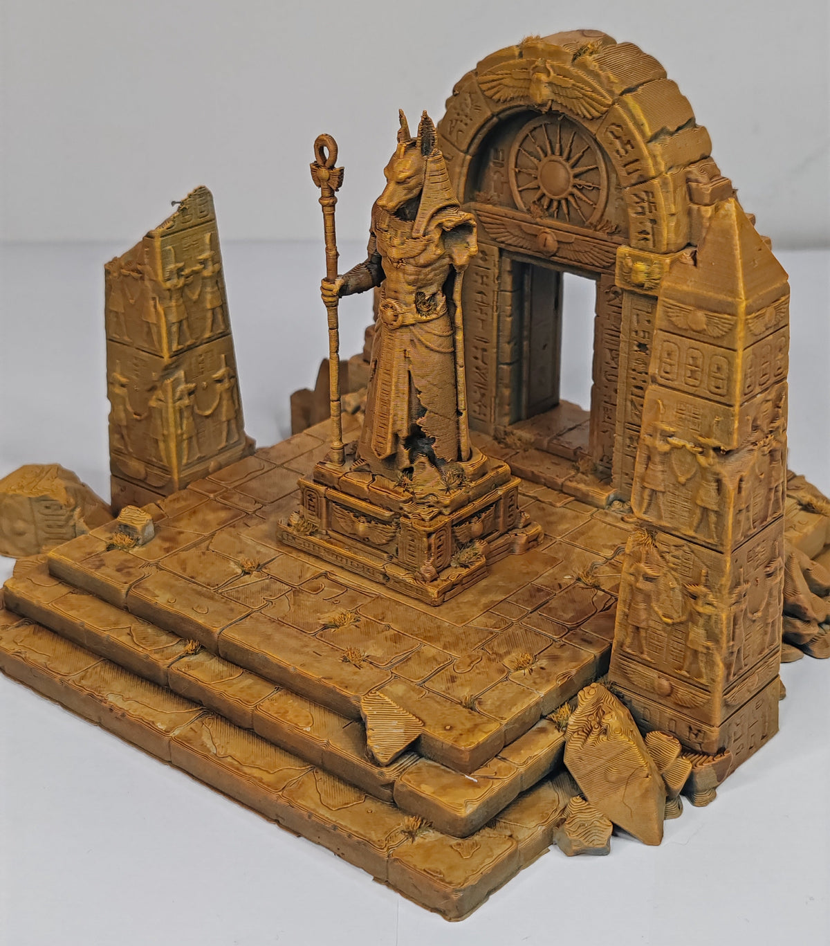 A detailed miniature model of an ancient stone temple or ruins set, featuring a central statue of a robed figure holding a staff, with carved architectural details, stairs, and debris on a plain light background.