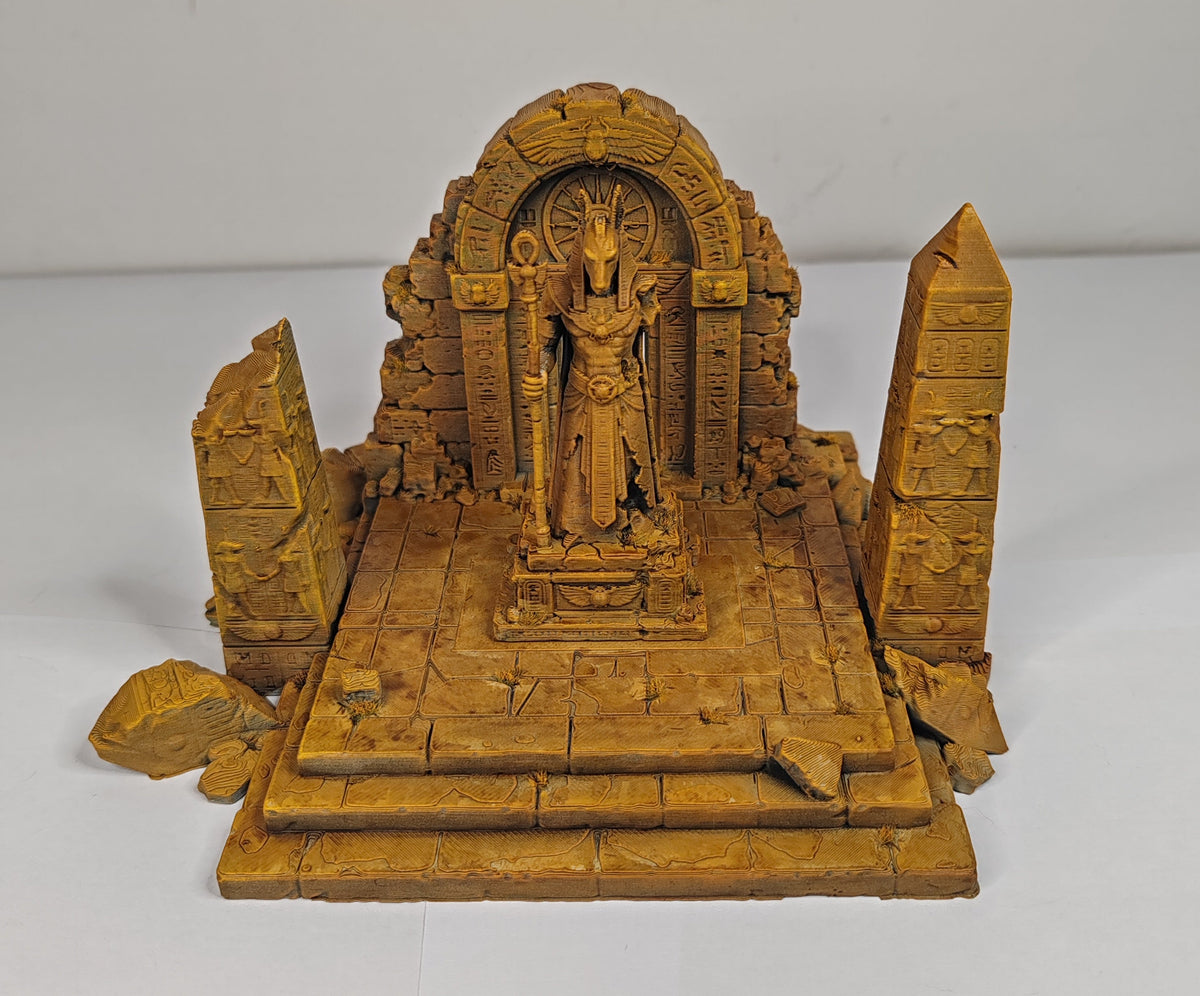 A small carved golden-brown miniature temple sculpture featuring a central deity statue, an arched niche, and flanking obelisks on a tiered platform, photographed against a plain light gray background.