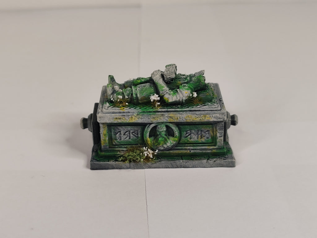 A miniature fantasy tombstone figurine with a green and gray stone texture, mossy details, and a small figure lying on top, photographed on a plain light background.