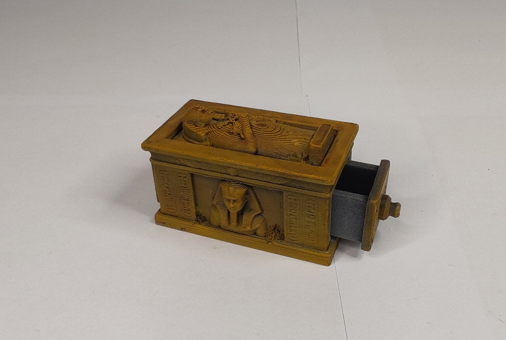 A small carved wooden Egyptian sarcophagus-shaped box with a partially open drawer, shown on a plain white surface.
