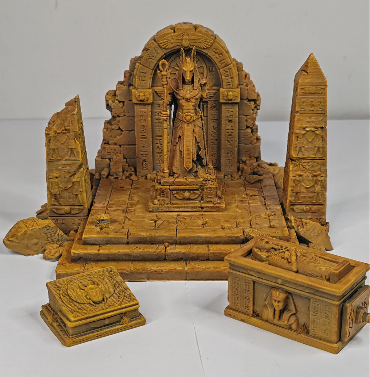 A set of miniature Egyptian-themed toy figurines and structures, including a central Anubis statue in an ancient temple, a sphinx, an obelisk, and a sarcophagus, all in a uniform terracotta clay color.